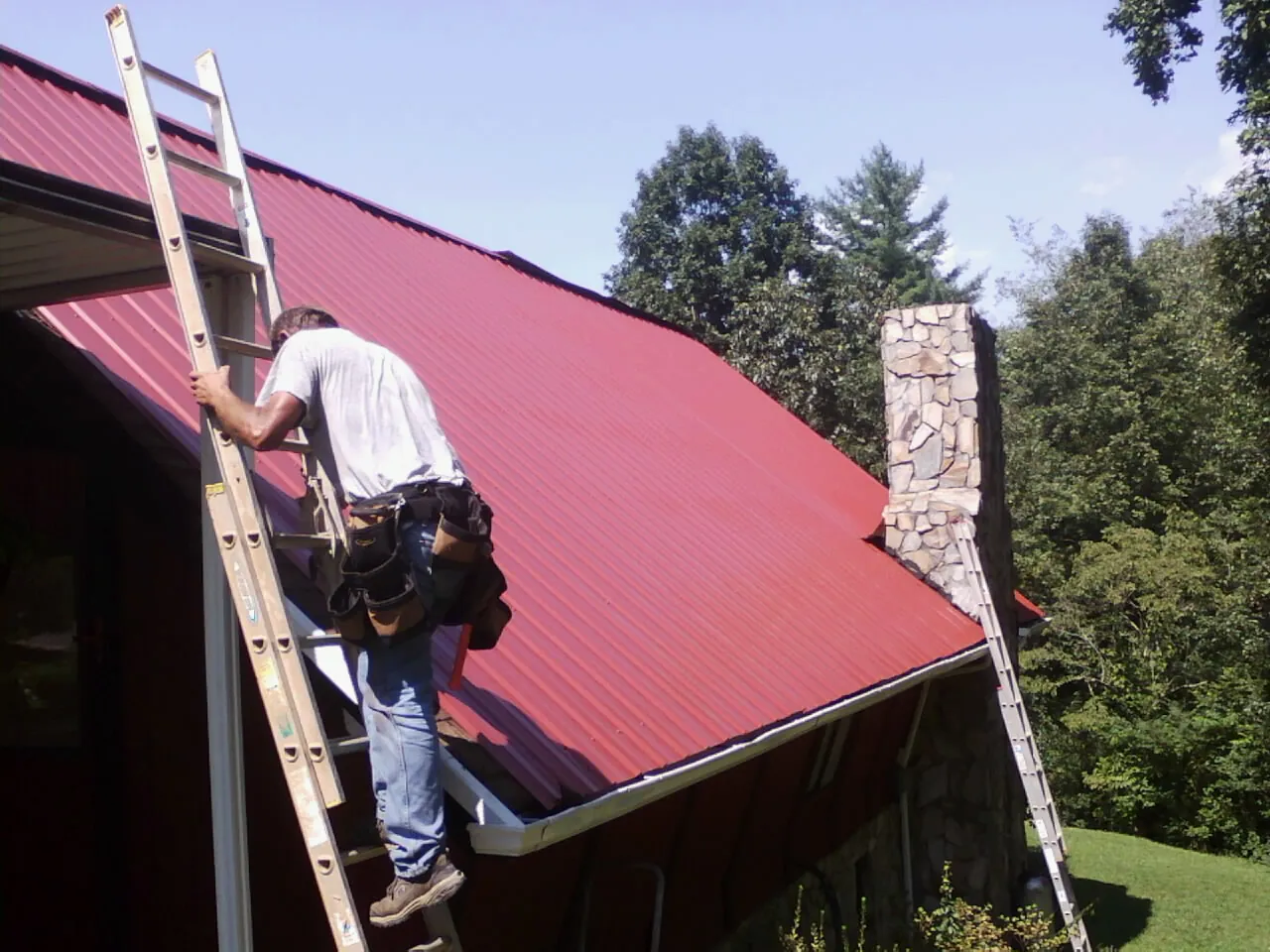Roofer on a ladder beside a red metal roof and stone chimney — Soffit & Fascia Repair by Roofing Brothers in La Vista